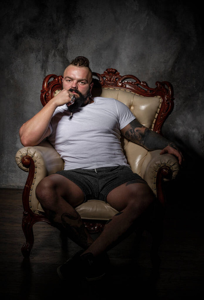 βάναυσος άντρας με μούσι και τατουάζ κάθεται σε μια καρέκλα. χτενισμένο ουίσκι και dreadlocks - Φωτογραφία, εικόνα