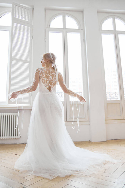 Beautiful slender blonde in the evening sun in a long white dress near a large window. Perfect hair and makeup for the bride, a new collection of wedding dresses - Valokuva, kuva