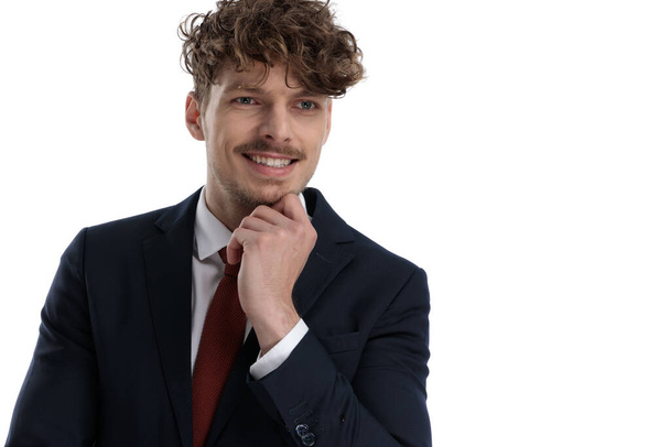 Γοητευτικός επιχειρηματίας που γελάει με το χέρι στο πηγούνι ενώ φοράει κοστούμι και στέκεται σε λευκό φόντο στούντιο - Φωτογραφία, εικόνα