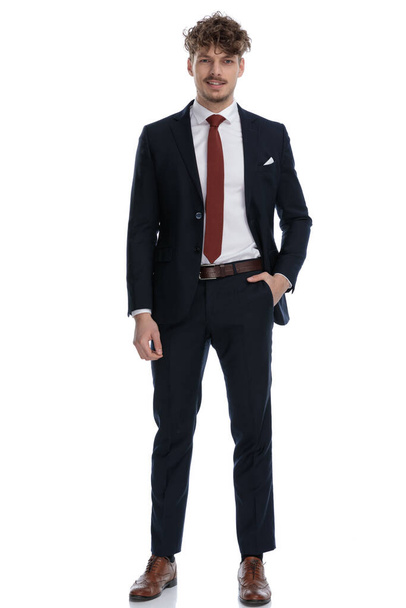 Positieve zakenman glimlachen met zijn hand in zijn zak tijdens het dragen van pak en staan op witte studio achtergrond - Foto, afbeelding