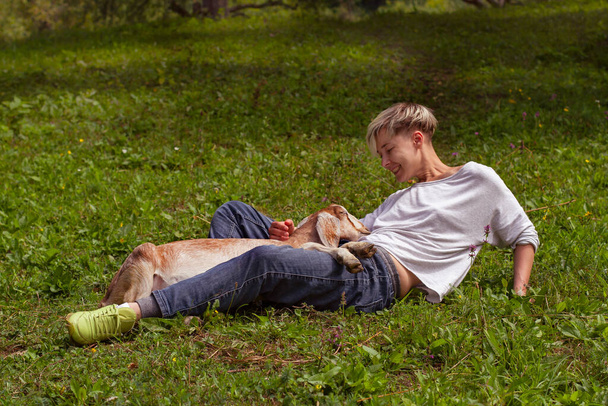 happy cheerful woman lies on the grass with a little goat. friendship of people and animals on a farm - Foto, Imagem
