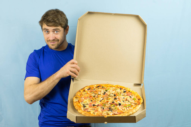 おいしいオープンボックスのピザを保持し、青い背景にクールな兆候を示しているハンサムな若い男。食べ物、ビジネスランチや配達サービスのために手にオープンピザボックスと青い背景に立つ男. - 写真・画像