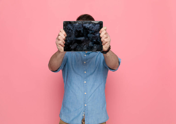 Casual Mann versteckt sein Gesicht vor der Kamera hinter seinem Tablet, trägt ein blaues Hemd und steht auf rosa Studiohintergrund - Foto, Bild