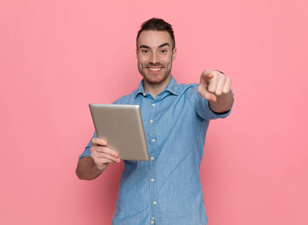mladý ležérní muž ukazuje dopředu, píše zprávu na tablet a usmívá se na růžové pozadí studia - Fotografie, Obrázek