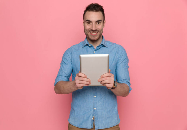 sexy casual man holding his tablet in front of the camera, smiling and wearing a blue shirt on pink studio background - Photo, Image