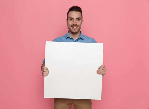 young handsome casual man holding a blank board and smiling on pink studio background - Fotoğraf, Görsel