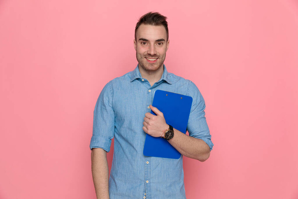 sexy hombre casual sosteniendo su portapapeles en el pecho, sonriendo y usando una camisa azul sobre fondo rosa estudio
 - Foto, Imagen