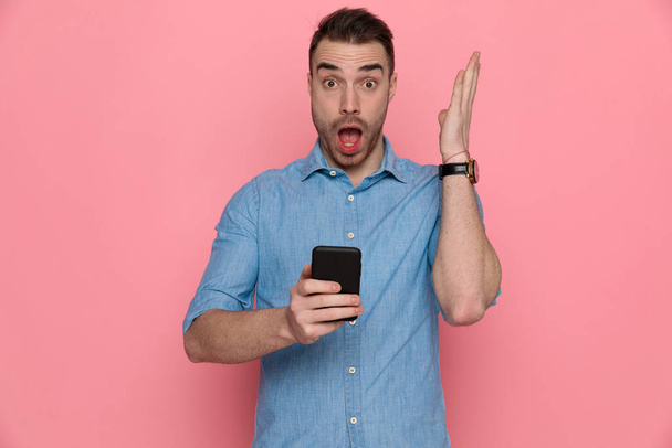 atraente chocado casual homem levantando uma mão e segurando seu telefone no fundo do estúdio rosa
 - Foto, Imagem