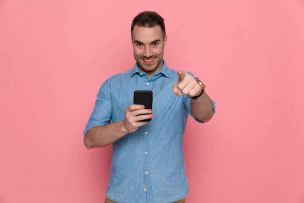 schöner, lässiger Mann, der nach vorne zeigt, eine Nachricht auf sein Handy schreibt und auf rosa Studiohintergrund lächelt - Foto, Bild