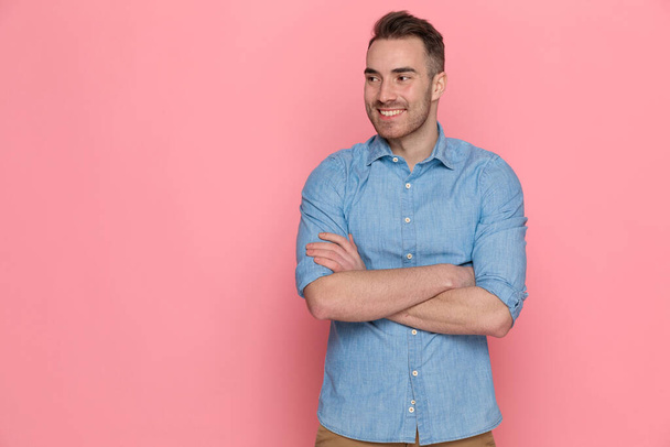 jonge knappe casual man met een blauw shirt, kruising van armen naar borst en opzij te kijken op roze studio achtergrond - Foto, afbeelding