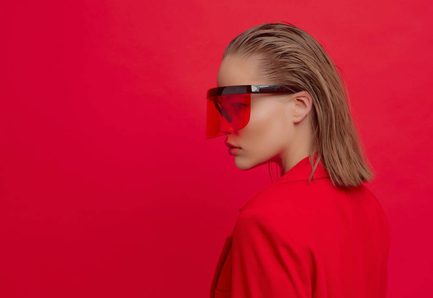 A cool stylish girl with a fashionable hairstyle and stylish glasses with a large glass poses on a bright red background - Фото, изображение