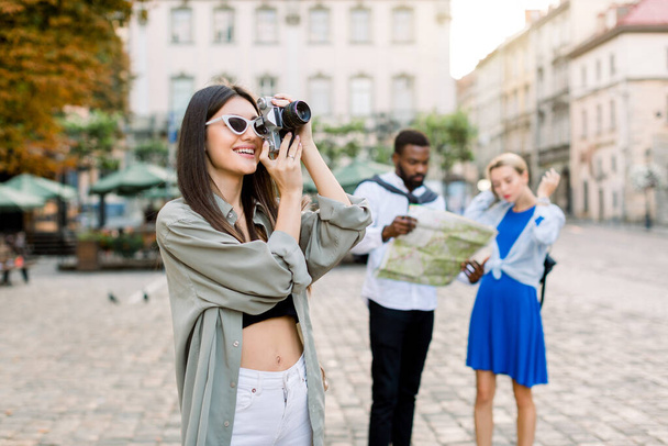 Pretty Caucasian brunette girl tourist in casual clothes taking photo in the city. African man and beautiful blond girl looking at the city map for some interesting places, standing on the background - Фото, изображение