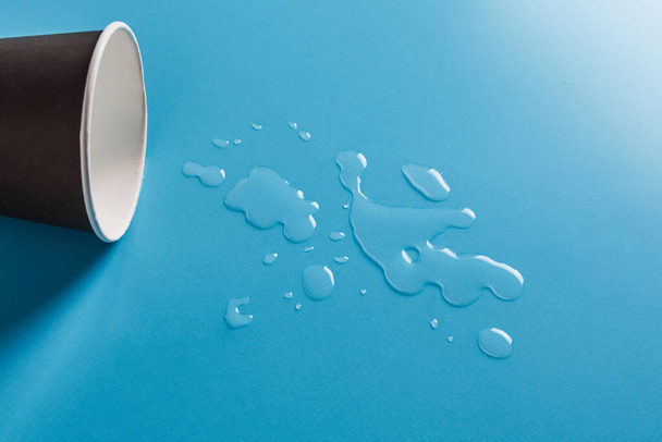 Dark Paper cup with abstract drops of spilled water on blue background. Spilled drink cup concept. - Фото, изображение