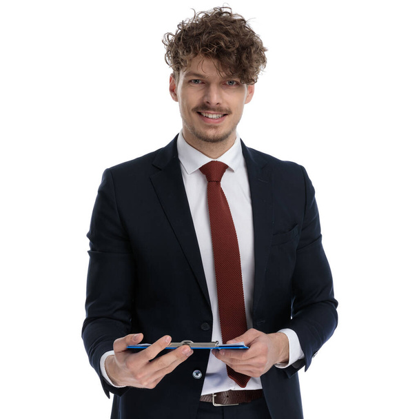 Empresário positivo segurando prancheta e sorrindo enquanto vestindo terno e de pé no fundo do estúdio branco
 - Foto, Imagem