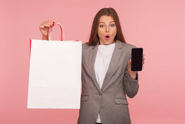 Wow, online shopping! Portrait of astonished elegant businesswoman in suit jacket holding bags with copy space for advertising text and showing cell phone, looking with surprised face. studio shot - Photo, Image