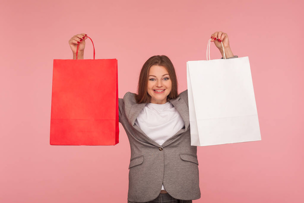 Portrait of attractive woman in business suit raising shopping bags and looking with joyful expression, smiling, satisfied with favorable purchase, discounts. studio shot isolated on pink background - Fotoğraf, Görsel