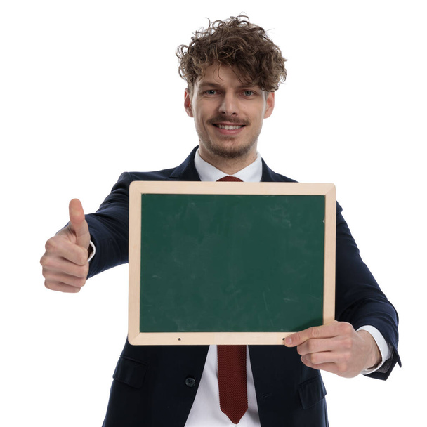 Happy businessman presenting green blackboard and gesturing ok while wearing suit and standing on white studio background - Foto, Bild