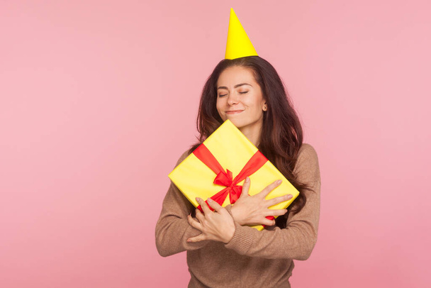 Portrait of amazing joyful satisfied woman with party cone embracing best holiday gift and closing eyes, smiling with pleased expression, celebrating birthday, anniversary. studio shot pink background - Photo, image