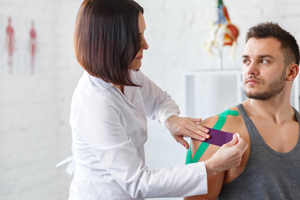 Kinesiology taping. Physical therapist applying kinesiology tape to patient shoulder. Female therapist treating injured shoulder of male athlete. Post traumatic rehabilitation, sport physical therapy - Foto, Bild