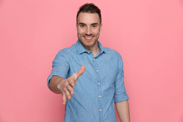 young attractive casual man extending his hand, ready for shaking hands on pink studio background - Zdjęcie, obraz
