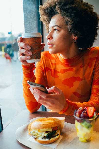 Multiethnic beautiful young woman sitting restaurant having lunch drinking reusable cup thinking using smartphone - healthy, technology, thinking concept - Foto, imagen