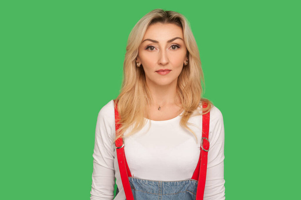 Portrait of serious concentrated adult woman with blond hair in stylish denim overalls looking at camera with focused calm pensive face expression. indoor studio shot isolated on green background - Φωτογραφία, εικόνα