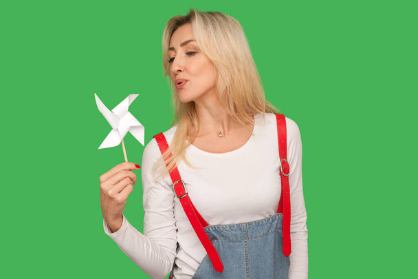 Pinwheel toy. Portrait of beautiful adult woman in stylish overalls blowing at paper windmill, playing with origami hand mill on stick, having fun. indoor studio shot isolated on green background - Photo, Image