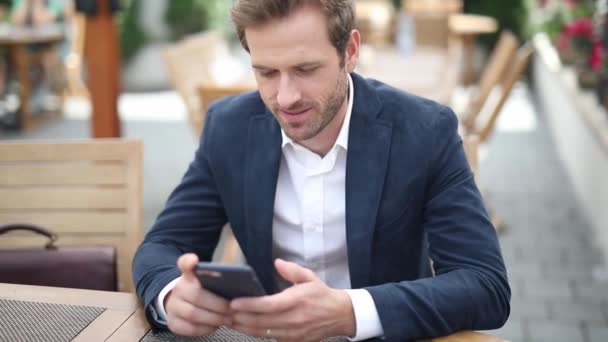 elegant young businessman sitting at a table, reading on his phone and smiling at camera - Filmmaterial, Video