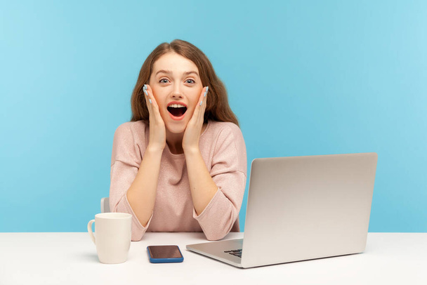 Wow, unbelievable! Surprised astonished young woman employee sitting at workplace with open mouth, shouting in amazement, shocked by unexpected news. indoor studio shot isolated on blue background - Foto, Bild