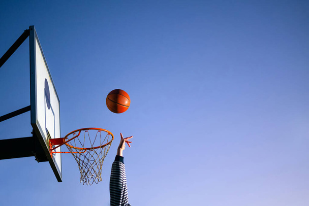 Street basketball ball player throwing ball into the hoop. Close up of hand, orange ball above the hoop net with blue sky in the background. Concept of success, scoring points and winning. Copy space - Photo, Image