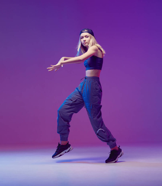 Cute teen girl dancing hip-hop in stylish clothes, a baseball cap, in a Studio with neon lighting. Dance color poster. - Foto, afbeelding