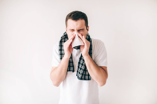 Young sick man isolated over white background. Guy sneezing into white napkin. Sick ill person with scarf around neck alone in studio. - Фото, изображение