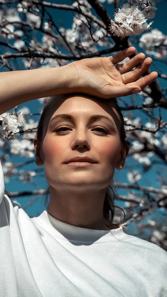 Closeup portrait of a beautiful girl on the background of blooming cherry tree, authentic beauty of young woman like a freshness of spring natur - Fotó, kép
