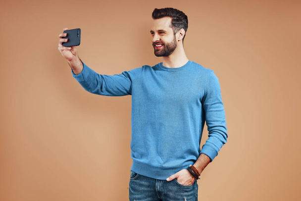 Hombre joven feliz en ropa casual inteligente usando tableta digital y tomando selfie mientras está de pie contra el fondo marrón
 - Foto, imagen