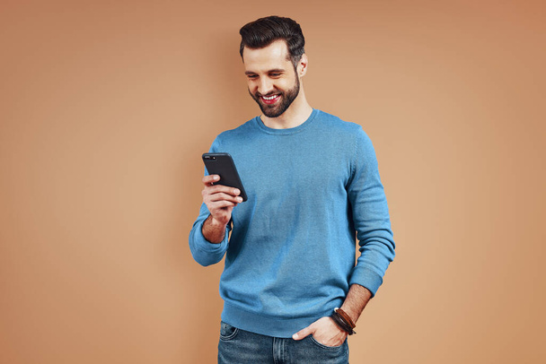 Hezký mladý muž v ležérní oblečení pomocí digitálního tabletu, zatímco stojí na hnědém pozadí - Fotografie, Obrázek