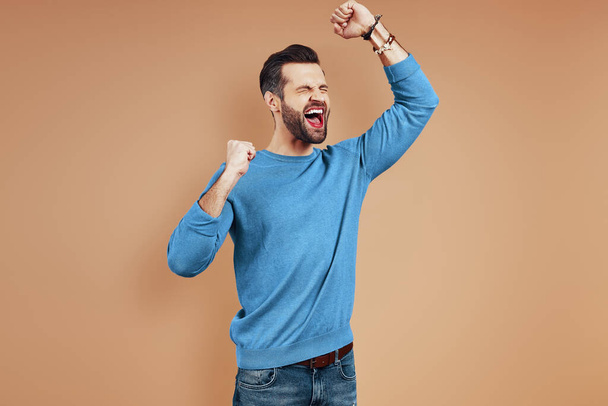 Cheerful young man in casual wear shouting and gesturing while standing against brown background - Foto, immagini