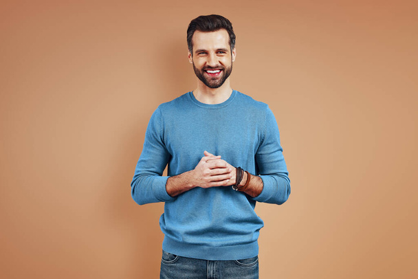 Charming young man in casual wear looking at camera and smiling while standing against brown background - Photo, Image