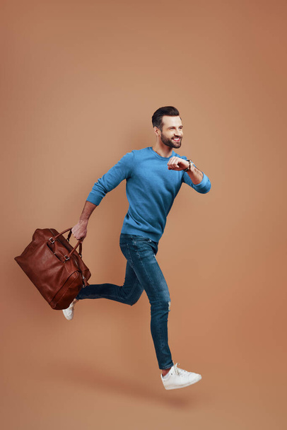 Full length of trendy young man with bag smiling while running against brown background - Foto, Imagen