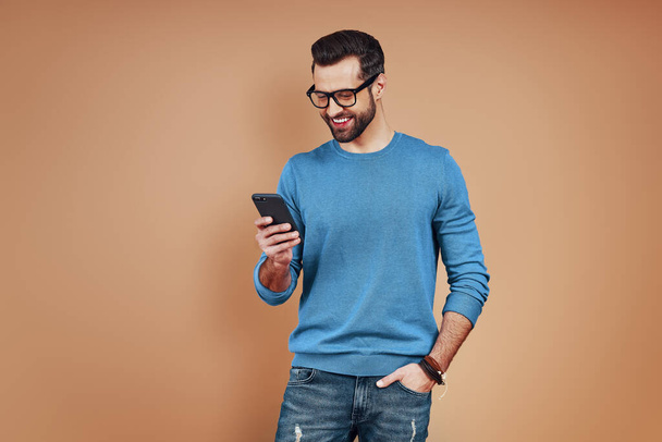 Akıllı gündelik giyinen mutlu genç adam akıllı telefon kullanıyor ve kahverengi arka planda dururken gülümsüyor. - Fotoğraf, Görsel
