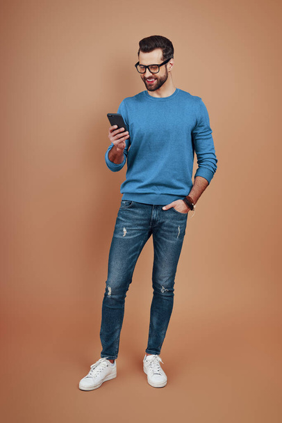 Teljes hossza boldog fiatalember okos alkalmi viselet segítségével okos telefon és mosolygós, miközben áll szemben a barna háttér           - Fotó, kép