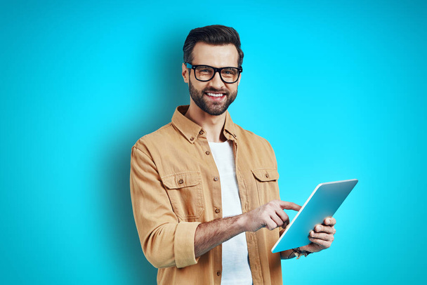Rušný mladý muž v inteligentní ležérní oblečení pomocí digitálního tabletu a při pohledu na fotoaparát, zatímco stojí na modrém pozadí - Fotografie, Obrázek