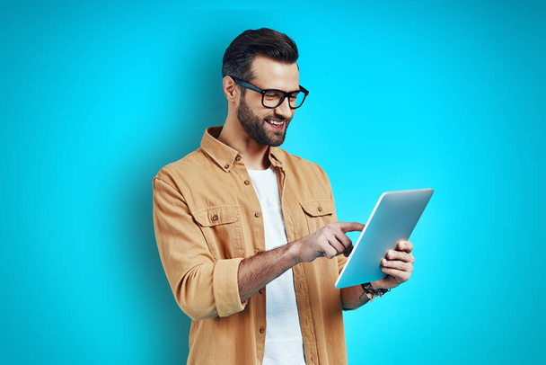 Mavi arka planda dikilirken dijital tablet kullanan akıllı ve rahat giyinen genç bir adam.       - Fotoğraf, Görsel