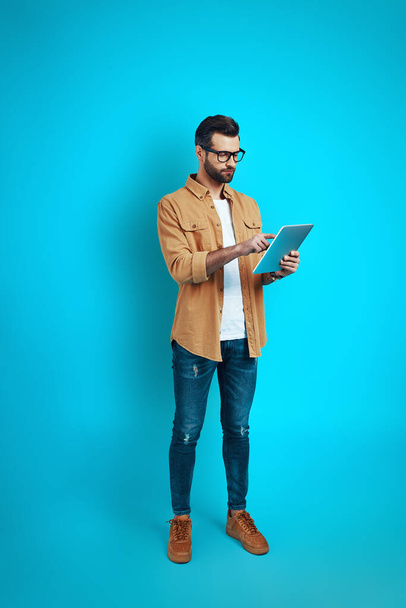 Volledige lengte van de jonge man in slimme casual slijtage met behulp van digitale tablet, terwijl staan tegen een blauwe achtergrond                  - Foto, afbeelding