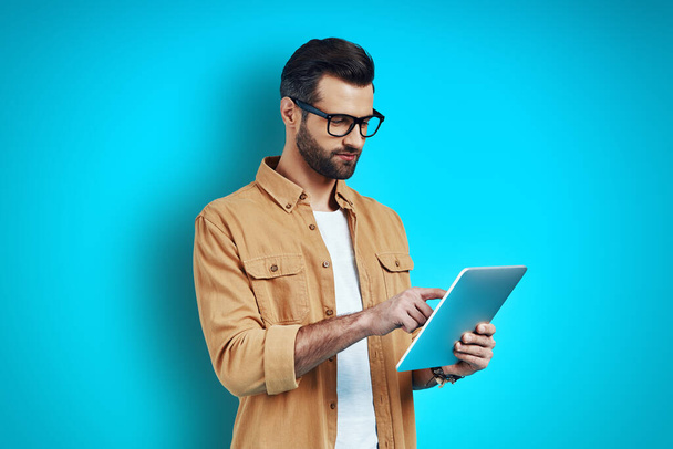 Hombre joven ocupado en ropa casual inteligente usando tableta digital mientras está de pie contra el fondo azul
 - Foto, Imagen
