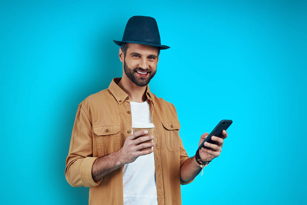 Модный молодой человек улыбается и использует смартфон, стоя на синем фоне
 - Фото, изображение