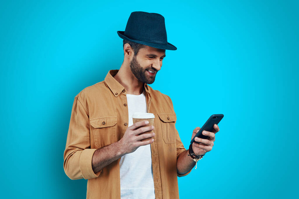 Jóképű fiatalember mosolyog és okos telefont használ, miközben a kék háttér előtt áll - Fotó, kép
