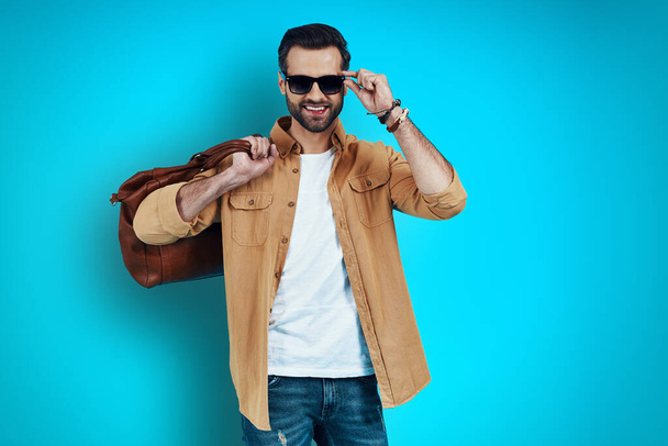 Charming young man smiling and adjusting eyewear while standing against blue background    - Photo, Image