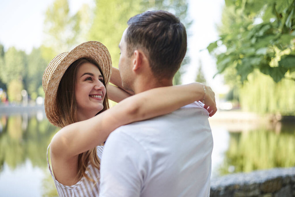 Young couple in love, hugging in the park in summer, smiling. Pretty blond girl in stripy overall and straw hat on romantic date with handsome brunette guy in white t-shirt. Sunday city walk. - Foto, afbeelding