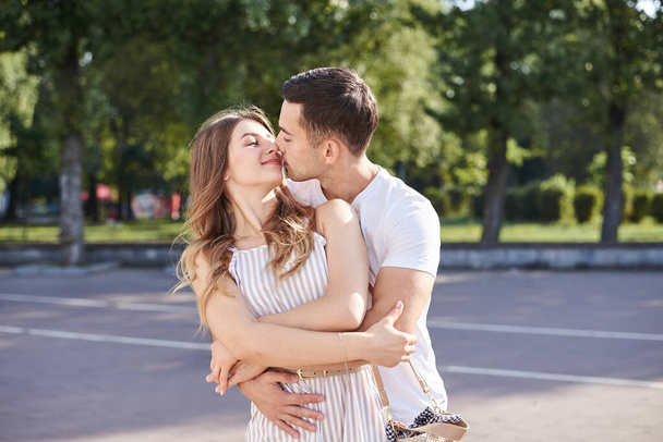 Handsome brunette guy, wearing white t-shirt, hugging and kissing pretty blond woman in stripy overall. Young couple in love, embracing in green park in summer. Romantic relationship. Valentines day. - Foto, imagen