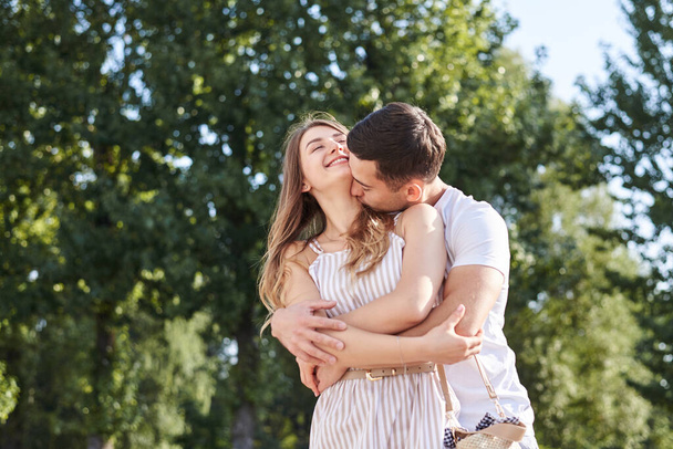 Handsome brunette guy, wearing white t-shirt, hugging and kissing pretty blond woman in stripy overall. Young couple in love, embracing in green park in summer. Romantic relationship. Valentines day. - Foto, afbeelding